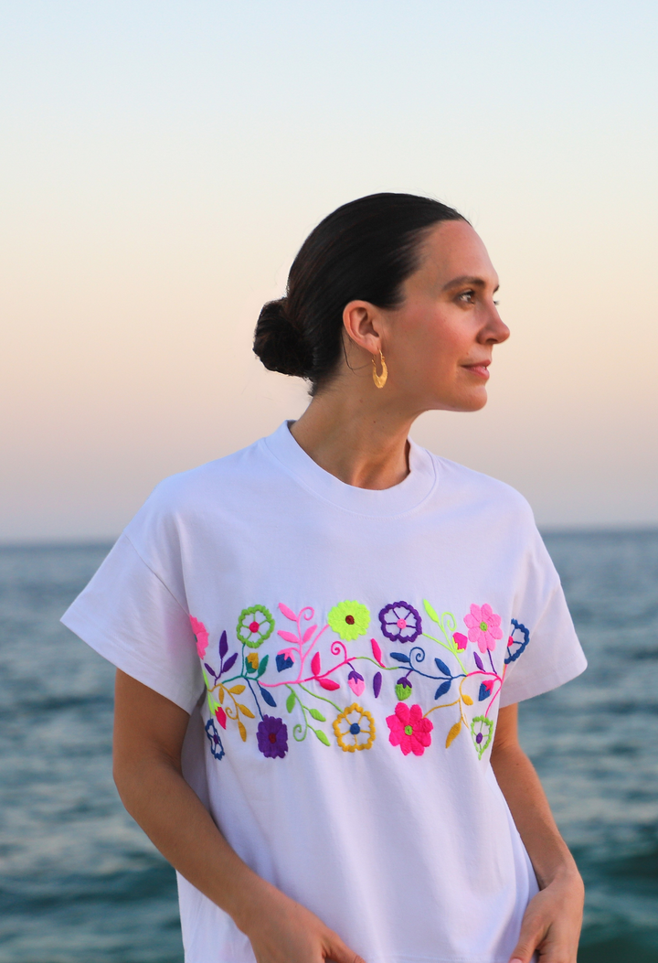 A woman standing by the sea, wearing a white short-sleeved t-shirt with vibrant floral embroidery across the chest.