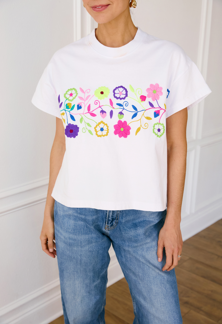 A person wearing a white crew neck t-shirt with colorful floral embroidery across the chest, paired with blue jeans.